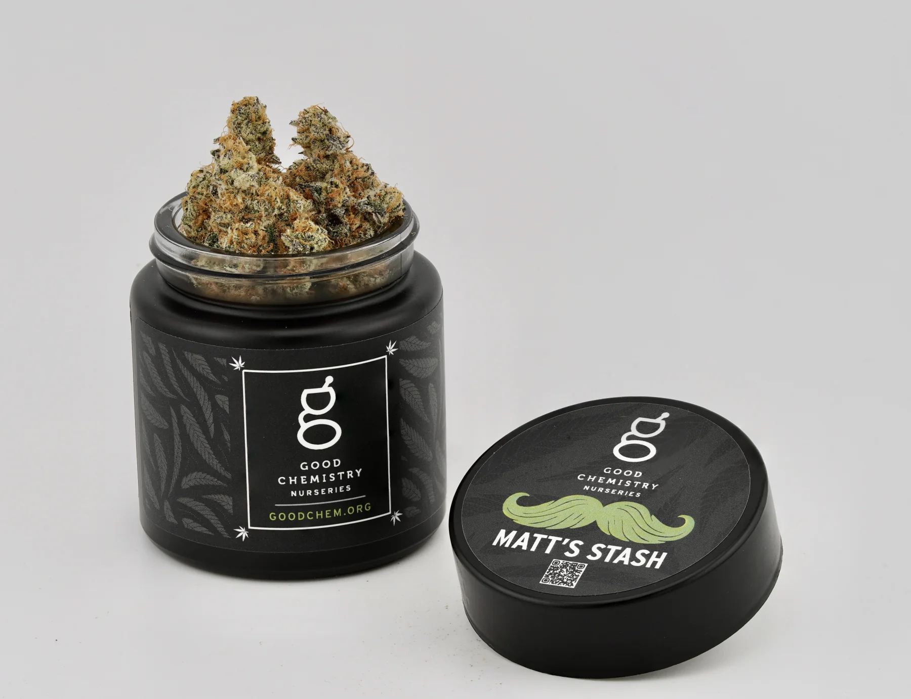 An open black Matt's Stash glass jar filled with cannabis flower beside its branded lid